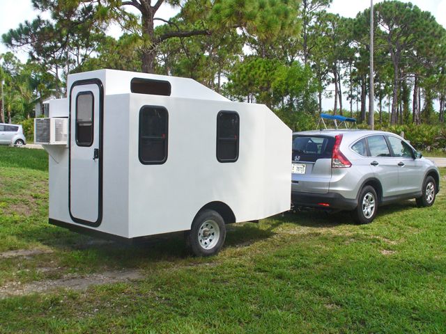 a small white trailer parked next to a car in a parking lot with trees and grass