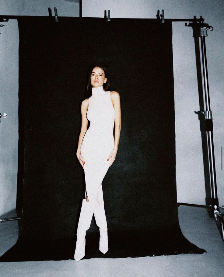 a woman standing in front of a black backdrop wearing white pants and knee high boots