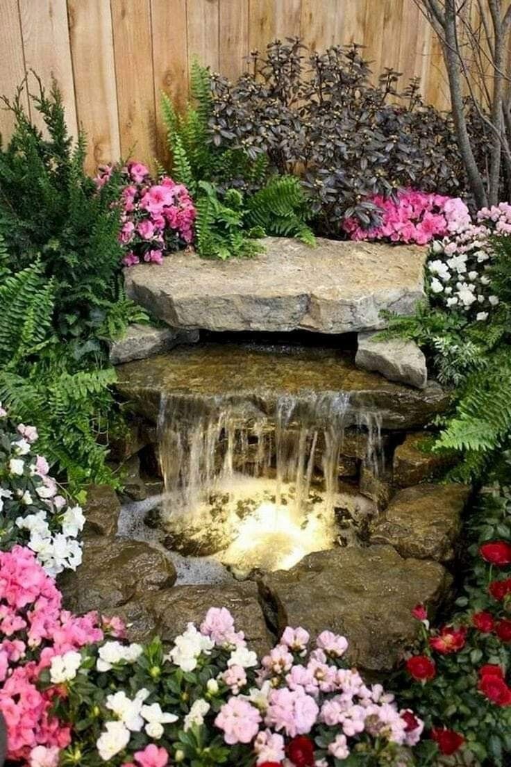 a garden with flowers, rocks and water features an artificial waterfall in the center surrounded by greenery