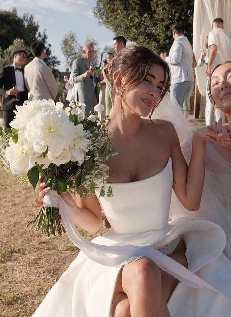 two brides sitting on the ground with flowers in their hands and people standing behind them