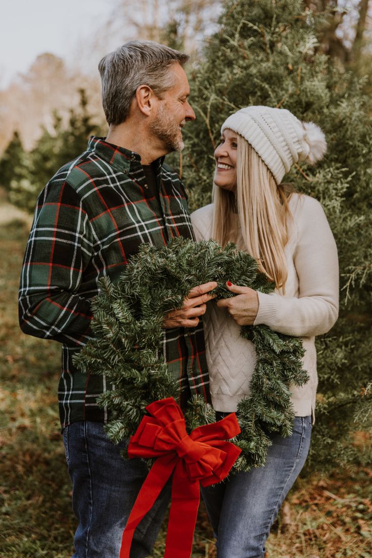 a man and woman standing next to each other in front of a christmas tree holding a wreath