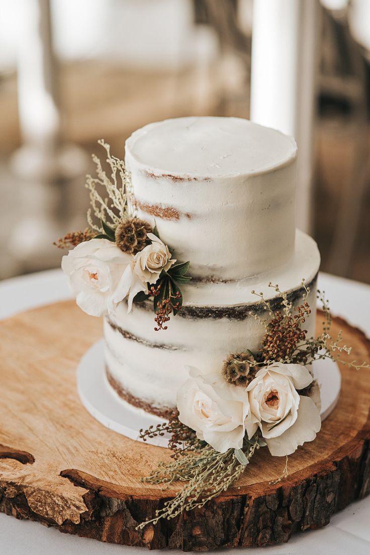 a wedding cake with white flowers and greenery sits on a wooden slice in front of the camera