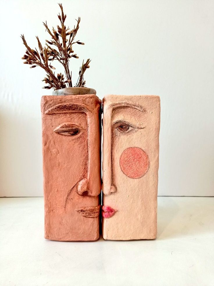 two clay blocks with faces on them and one has a plant in the vase next to it