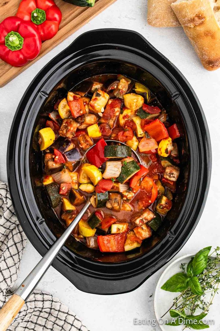 a slow cooker filled with vegetable stew next to sliced bread and fresh basil on the side