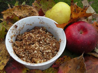 two apples and some granola in a white bowl on top of leaves with an apple next to it