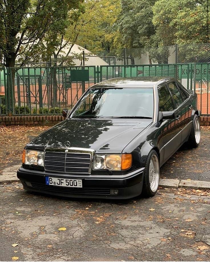 a black mercedes parked in front of a green fenced area with trees behind it