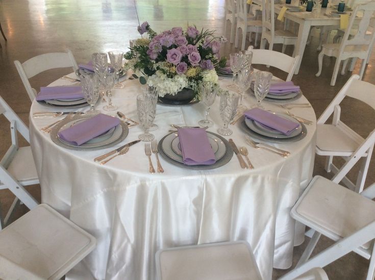 the table is set with white chairs and purple napkins, silverware, and flowers