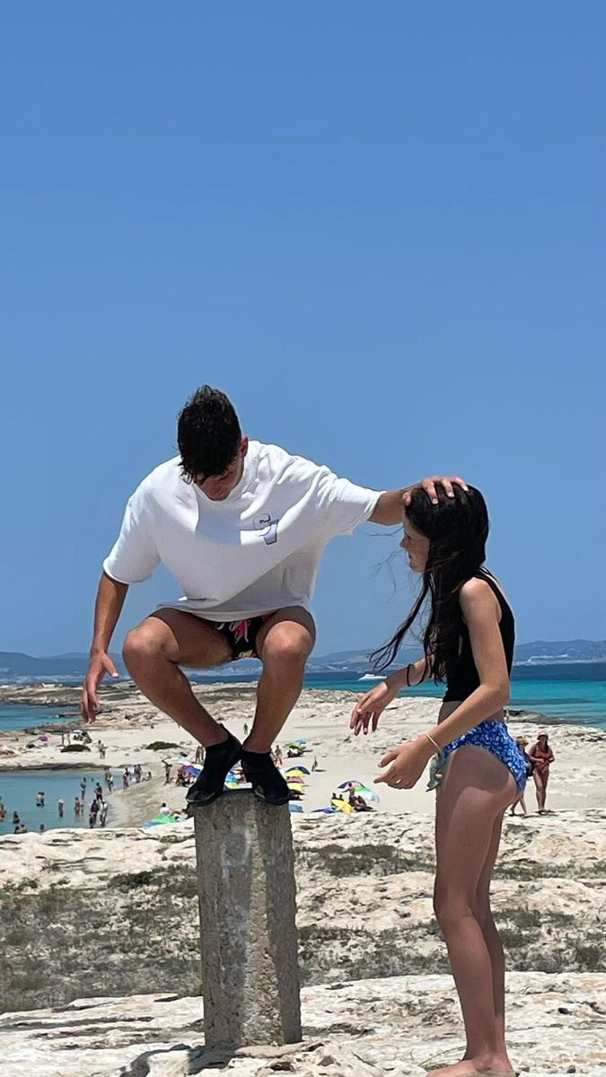 a man and woman standing on top of a stone pillar near the ocean with people swimming in the water