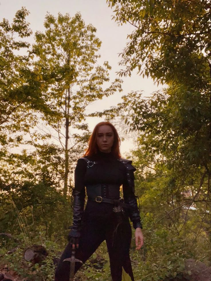 a woman with red hair and black leather outfit standing in the middle of a forest