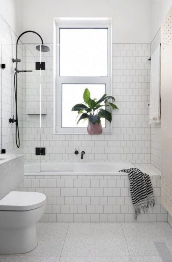 a white tiled bathroom with a toilet, bathtub and window in the corner that has a potted plant next to it