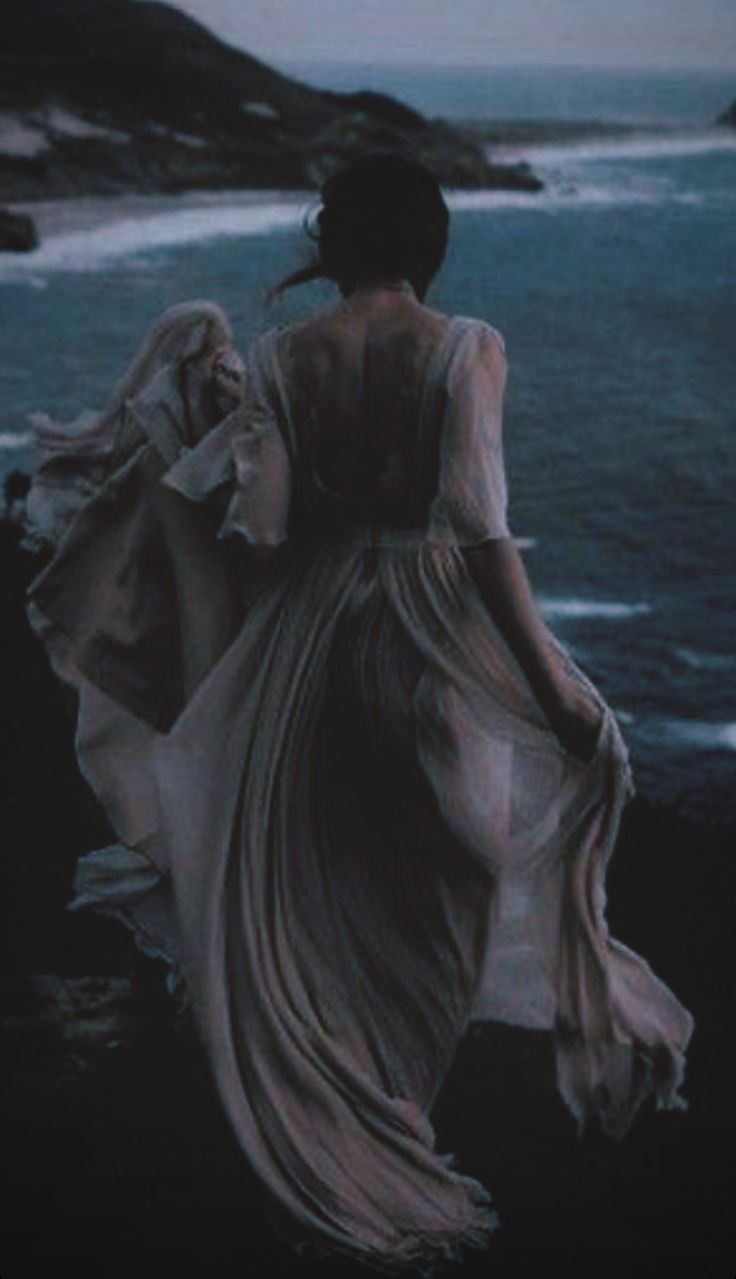 a woman in a long dress is walking by the ocean with her back to the camera