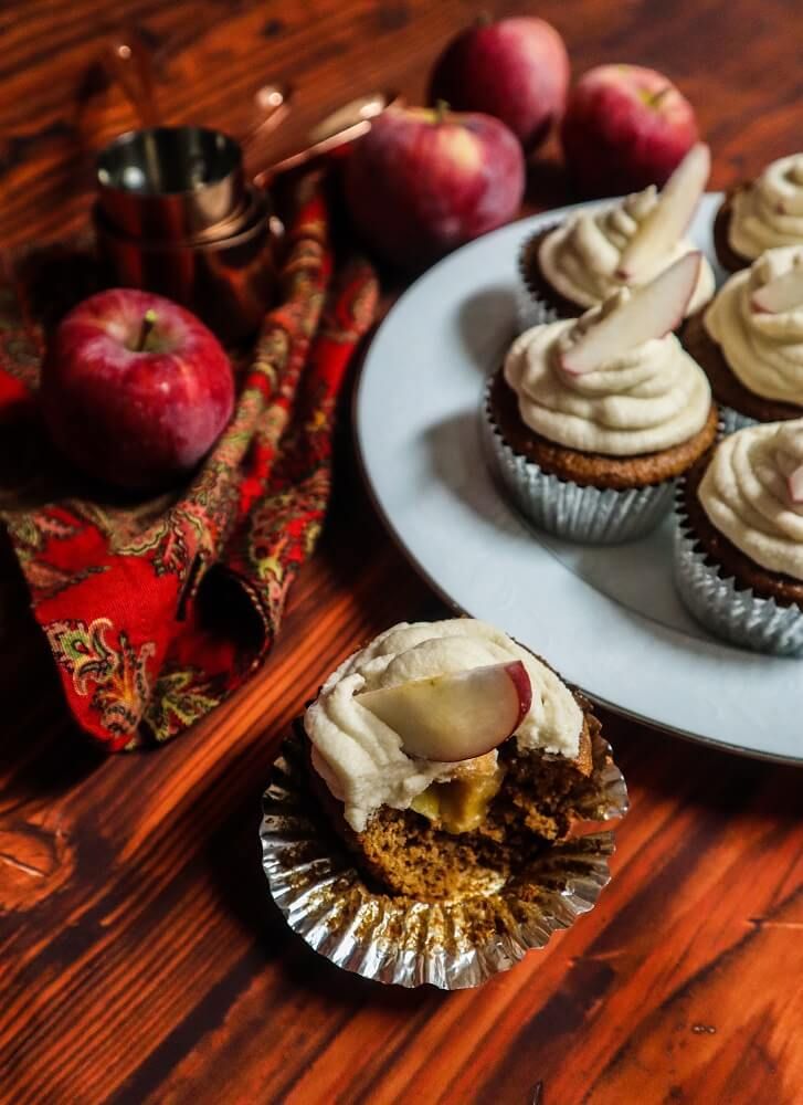 some cupcakes with icing and apples in the background on a wooden table