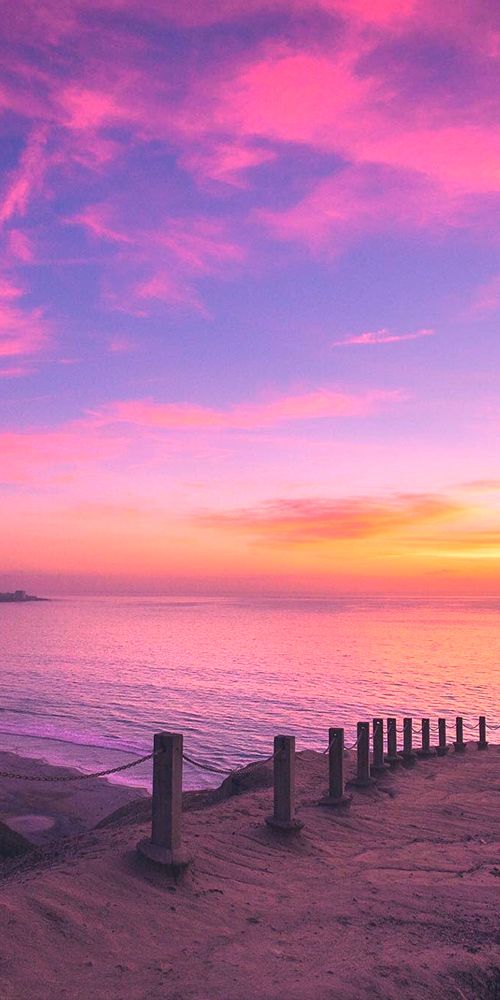 a bench sitting on top of a sandy beach next to the ocean under a purple and pink sky