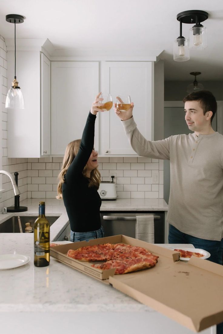 a man and woman toasting with pizza on the kitchen counter