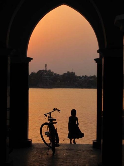 a woman standing next to a bike in front of a body of water at sunset