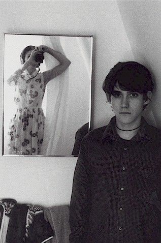 a young man standing in front of a mirror next to a pair of shoes on the floor