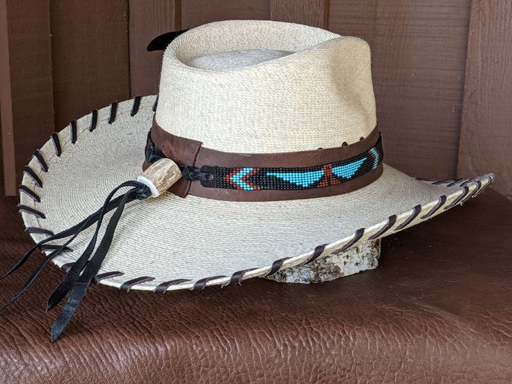 The Maverick starts with a Mexican soft palm leaf hat body that has a 3 3/4" brim and 5 1/4" crown. Hand laced in chocolate brown leather lace. The beaded hatband has a Thunderbird pattern and then mounted on distressed leather. The back of the band has a deer antler bead for accent. Starburst turquoise Concho and deep blue/green decorative feather completes the hat. All hats are made to order. Allow up to 4 weeks for your hat to ship. Brown Toquilla Straw Hat Bands For Ranch, Western Style Brown Panama Hat With Flat Crown, Brown Toquilla Straw Hat Band For Western-themed Events, Western Brown Panama Hat With Flat Crown, Rustic Natural Hat Bands For Western-themed Events, Handmade Fitted Brown Hat Bands, Brown High Crown Hat For Western-themed Events, Traditional Brimmed Straw Hat For Rodeo, Artisan Leather Hat For Rodeo