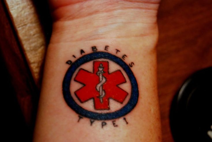 a small red and blue medical symbol tattoo on the left inner wrist, with words written below it