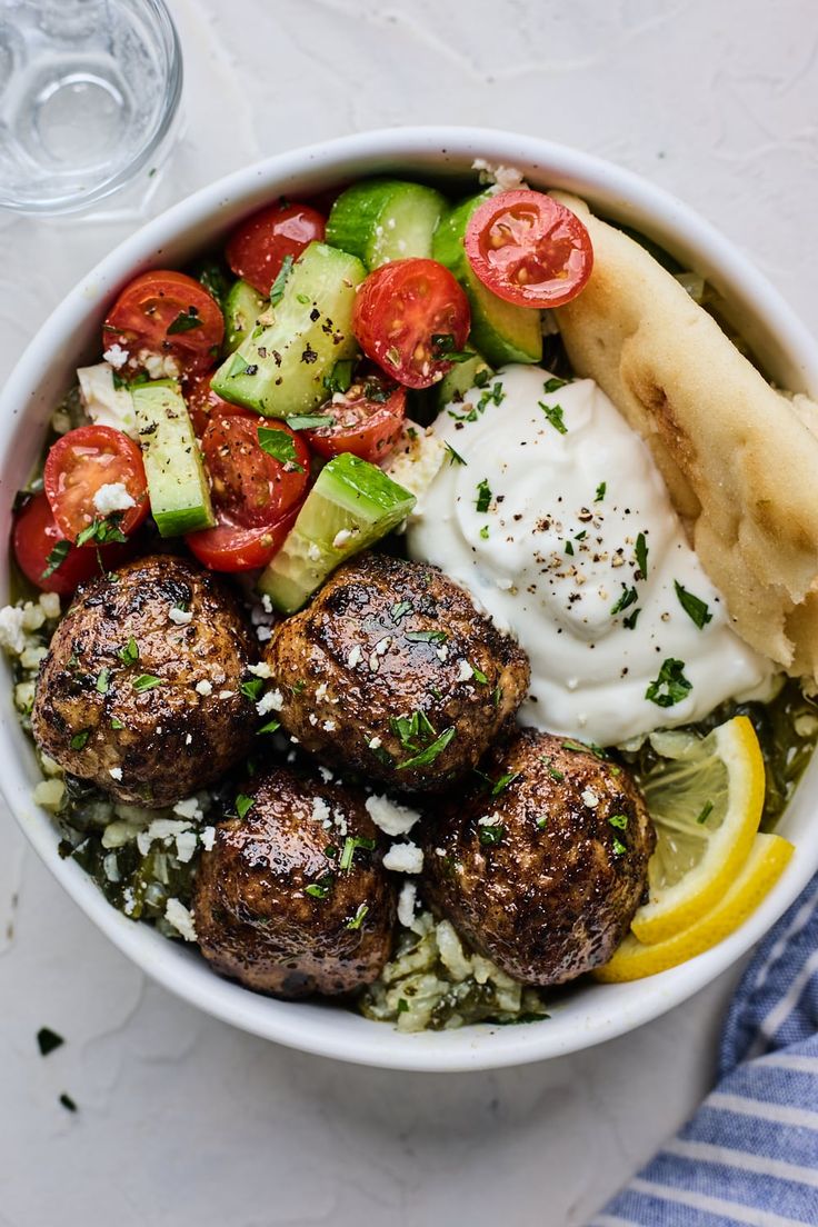 a white bowl filled with meatballs, cucumber, tomatoes and other veggies