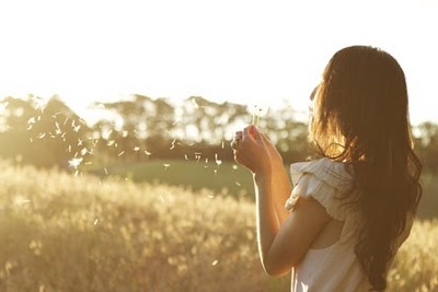 a woman standing in a field blowing on dandelions with the sun shining through her hair