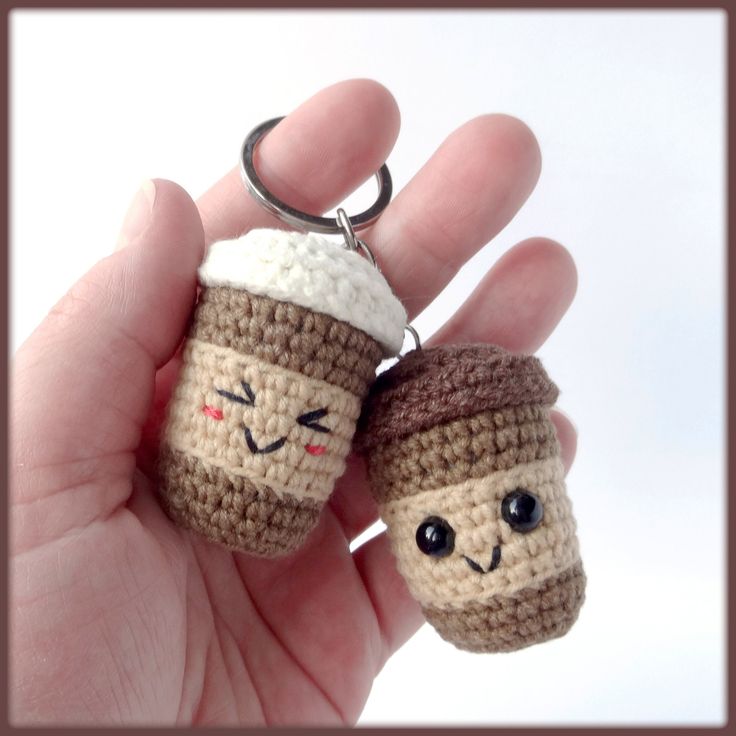 two crocheted coffee cup keychains with faces on them are held in the palm of a person's hand