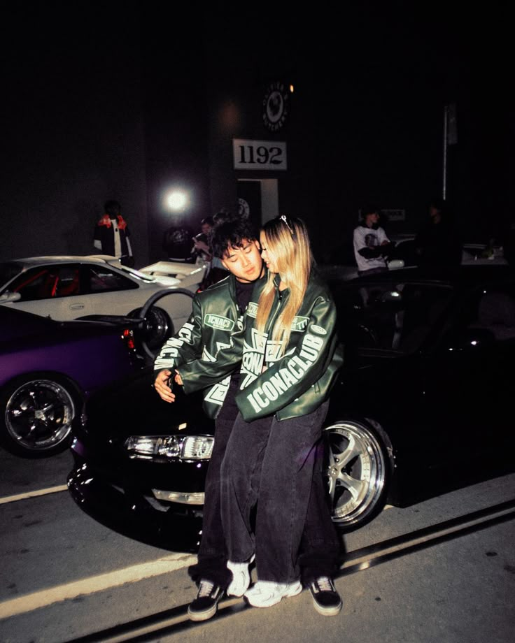 a man and woman sitting on the hood of a car in front of a gas station