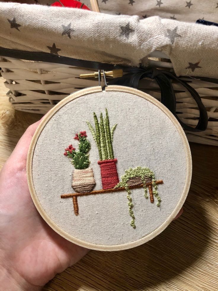 a hand holding a small embroideryed potted plant on a table next to a wicker basket