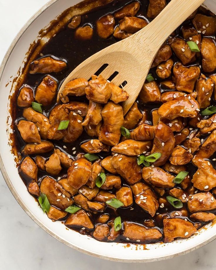 a white bowl filled with chicken and sauce on top of a table next to a wooden spoon