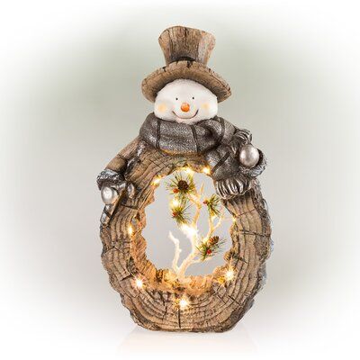 a snowman sitting on top of a tree stump holding a wreath with lights in it