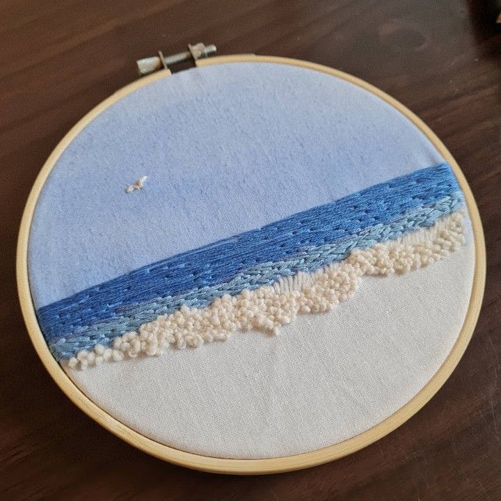 an embroidery project with blue and white waves on the beach in front of a wooden table