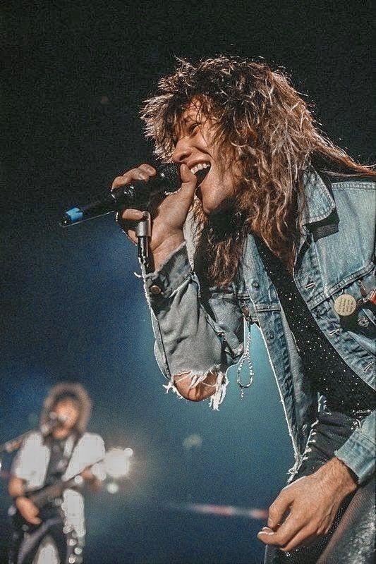 a man with long hair holding a microphone in front of another man singing on stage