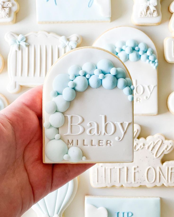 a hand holding a cookie with blue and white icing on it that says baby miller