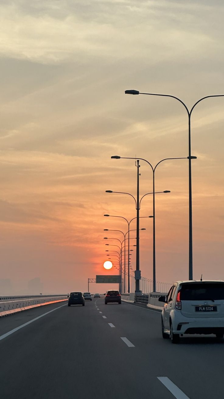 the sun is setting over the ocean and cars are driving down the road in front of it