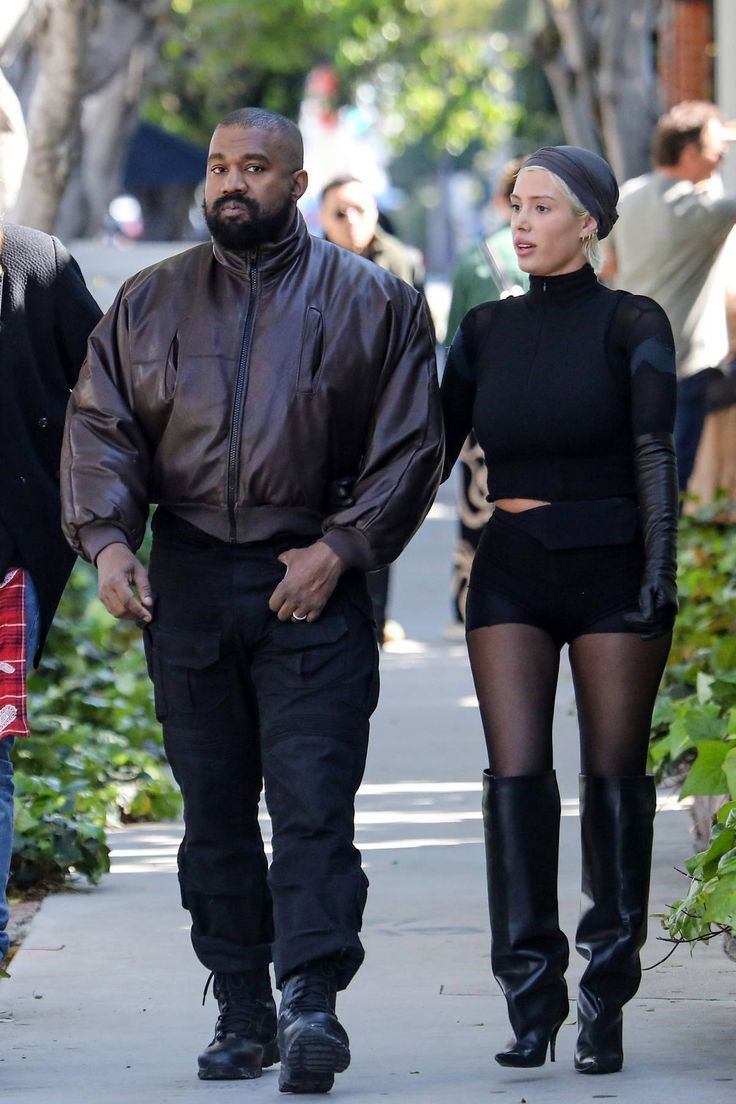 two women and a man walking down the street together, one in black is wearing thigh high boots