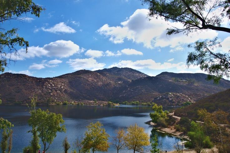 a lake surrounded by trees and mountains under a blue sky with white clouds in the background