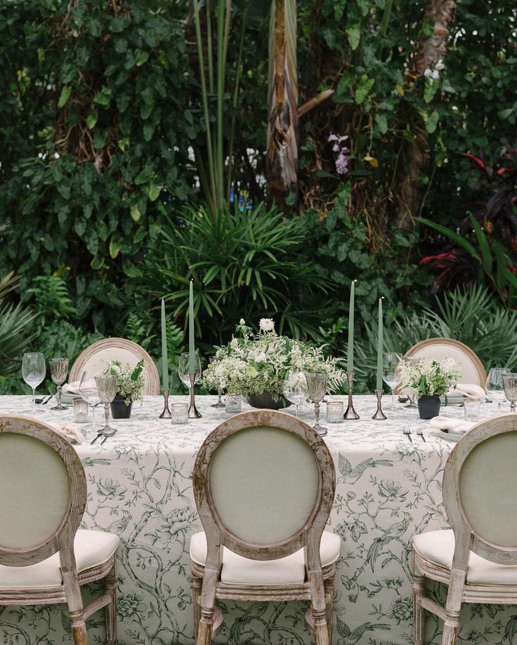 the table is set with white linens and chairs, surrounded by lush greenery