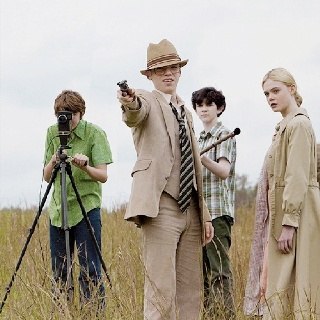 three people standing in tall grass with one holding a camera and the other pointing at something