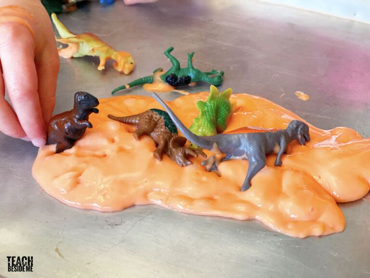 a hand reaching for toy dinosaurs on top of an orange substance