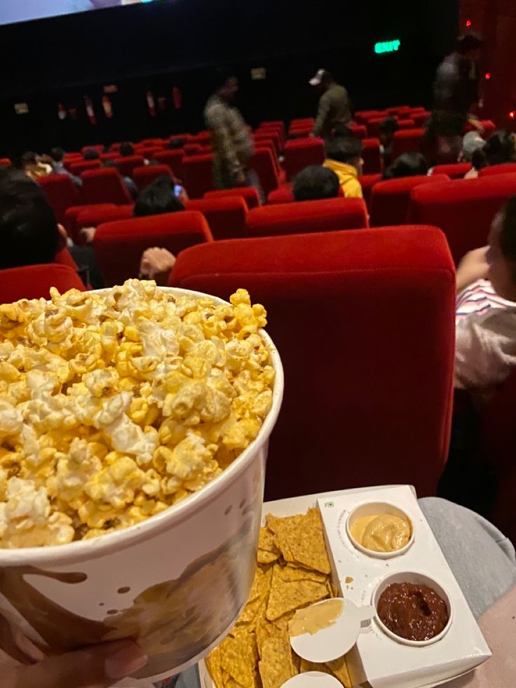 a person holding up a bowl of popcorn and chips in front of an audience at a movie theater