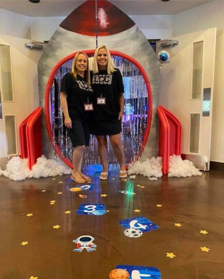 two women standing in front of a fake rocket ship with stars and confetti on the floor