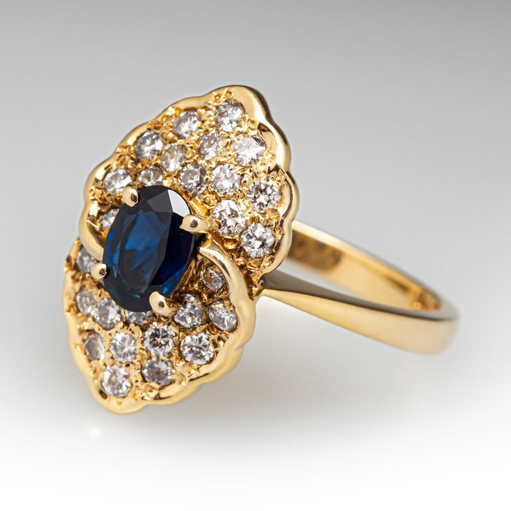 This pretty dinner ring is centered with one (1) oval cut natural sapphire set into a four-prong setting. The sapphire is bordered with twenty-eight (28) bead set round brilliant cut diamonds. The ring measures 18.1mm at the top and rises 8.3mm above the finger, tapering to 2.3mm wide and 1.2mm thick at the base of the shank. It is currently a size 6.5 and is crafted in 14k yellow gold. Classic Oval Sapphire Ring With Diamond Accents, Elegant Gold Ring With Lab-created Sapphire, Elegant Yellow Gold Lab-created Sapphire Ring, Classic Cluster Sapphire Diamond Ring, Classic Sapphire Cluster Diamond Ring, Classic Oval Sapphire Ring With Rose Cut Diamonds, Classic Marquise Sapphire Ring With Center Stone, Elegant Yellow Gold Rings With Lab-created Sapphire, Elegant Lab-created Sapphire Ring