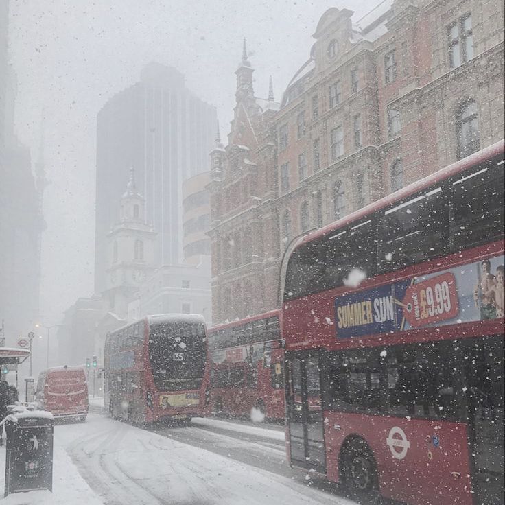 two red double decker buses driving down a snow covered street with tall buildings in the background