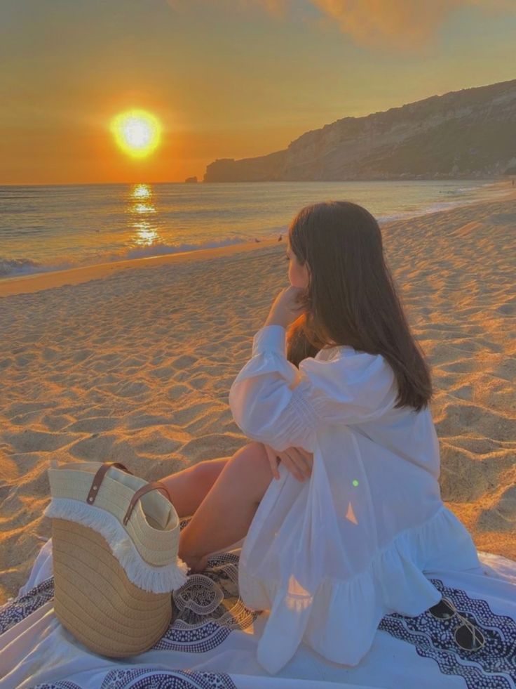 a woman sitting on top of a beach next to the ocean with a straw bag