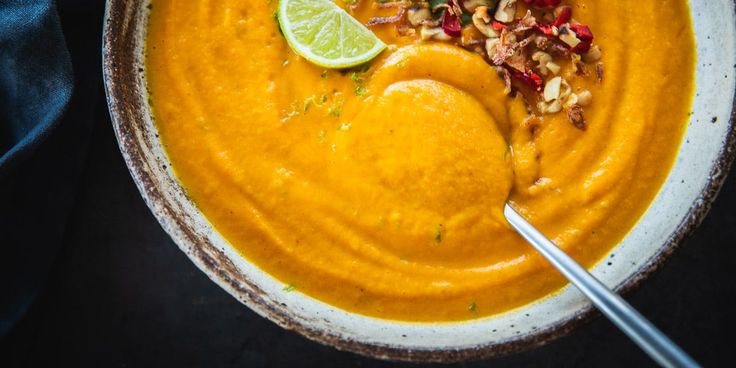 a white bowl filled with carrot soup and topped with chopped nuts, lime wedges and a spoon