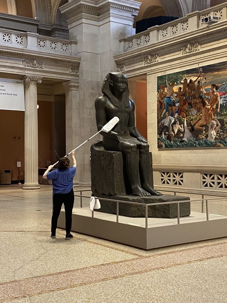 a woman holding a baseball bat standing next to a statue in a room with paintings on the walls