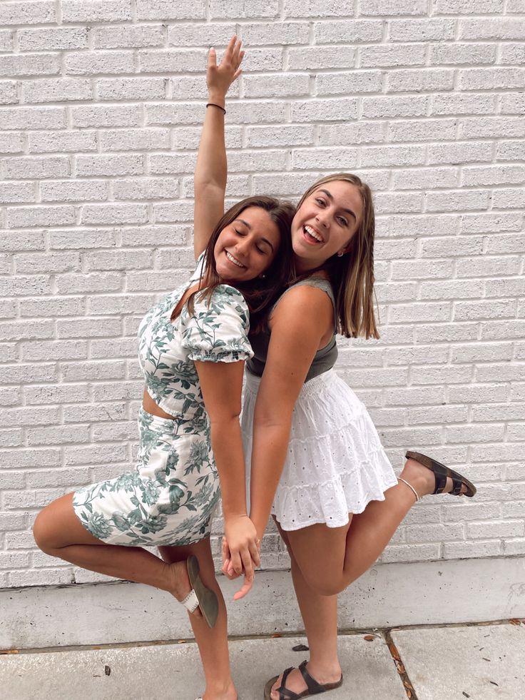 two young women posing for the camera in front of a white brick wall with their arms around each other