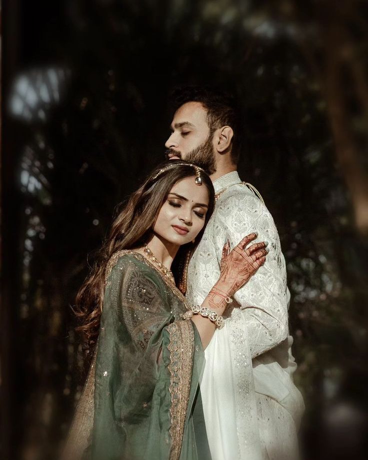 a man and woman standing next to each other in front of some trees with their arms around each other