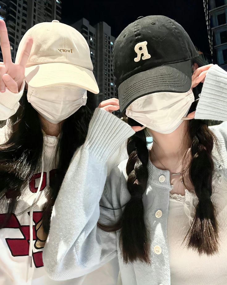 two girls wearing face masks and hats making the peace sign with their hands while standing next to each other