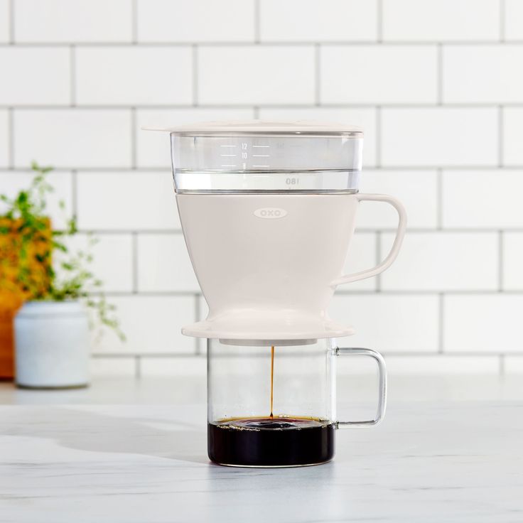 a glass coffee maker filled with liquid on top of a white counter next to a potted plant