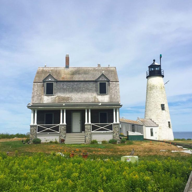 an old house with a lighthouse in the back ground and green bushes on either side